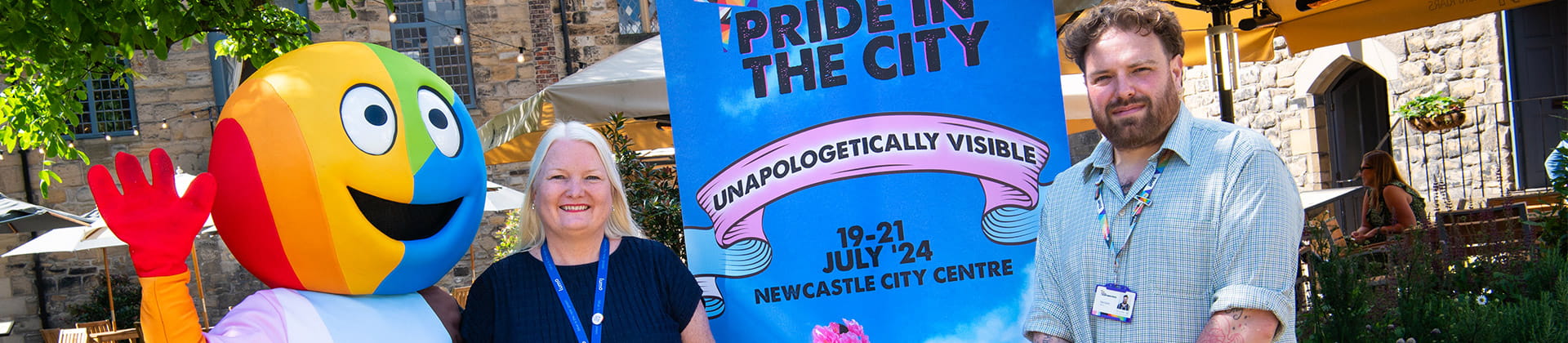 Two Lumo staff members standing with the pride mascot next to a pride in the city poster